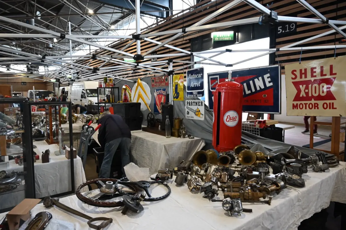 Stand de pièces détachées anciennes sur tables nappées de blanc, enseignes de pétroliers et pompe à essence d’époque à Epoqu’Auto