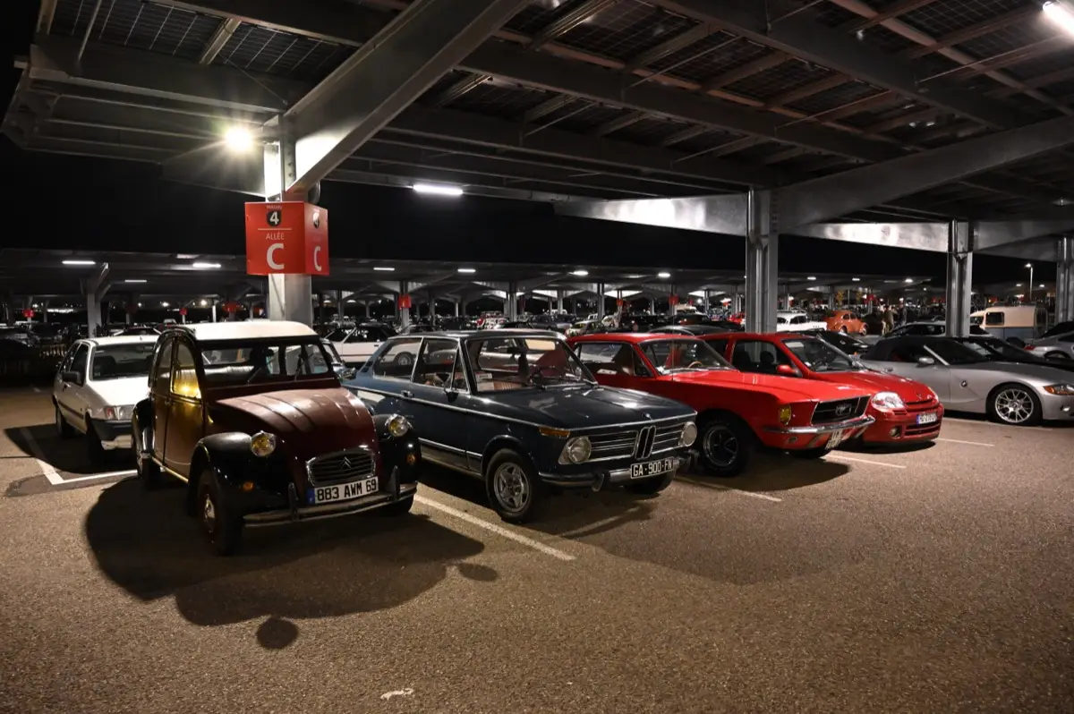 Citroën 2 CV, BMW et autres voitures anciennes de visiteurs garées sous l’auvent du parking P4 d’Eurexpo le soir