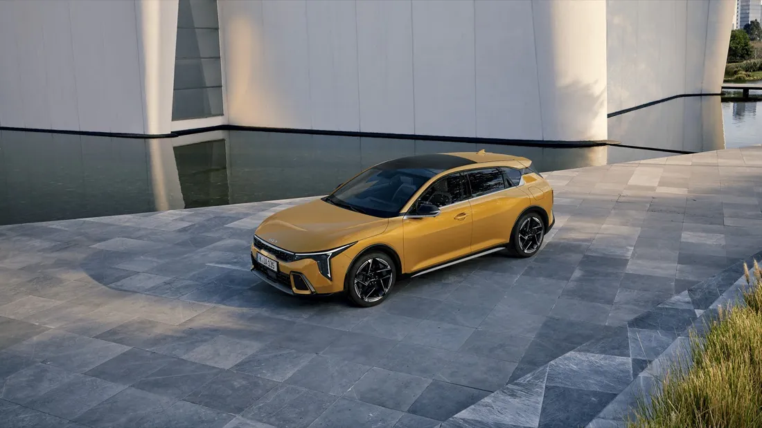 Kia K4 jaune garée sur une esplanade dallée au bord d’un bassin, vue de trois quarts avant