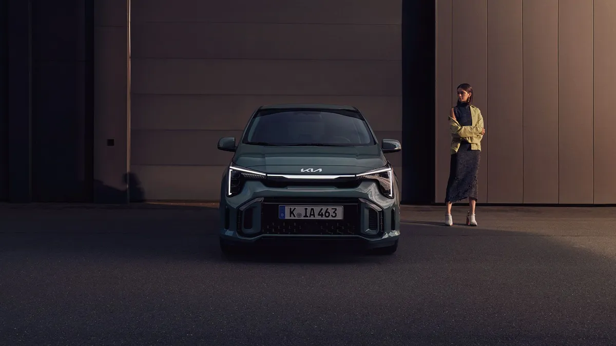 Kia Picanto restylée vert foncé vue de face devant un bâtiment, une jeune femme debout à côté